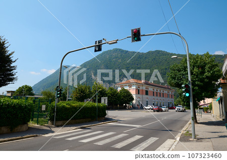 Pedestrian crossing, crosswalk and traffic light in the Italian town of Canso in Lombardy, with the Alps mountains in the background. Italy Pedestrian crossing, crosswalk and traffic light in the Italian town of Canso in Lombardy, with the Alps mountains in the background. Italy 107323460