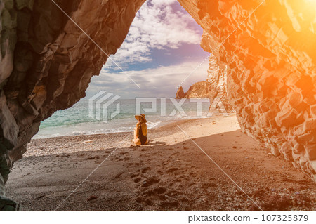 Woman travel sea. View of a woman in a black swimsuit from a sea cave Attractive woman enjoying the sea air sits on the beach and looks at the sea. Behind her are rocks and the sea 107325879