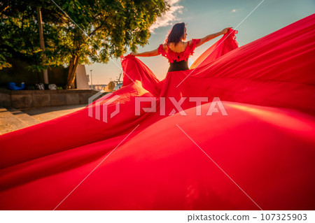Sunrise red dress. A woman in a long red dress against the backdrop of sunrise, bright golden light of the sun's rays. The concept of femininity, harmony. 107325903