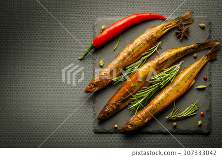 Smoked capelin fish on a serving board with rosemary and pepper served for lunch. Dark space for advertising Smoked capelin fish on a serving board with rosemary and pepper served for lunch. Dark space for advertising 107333042