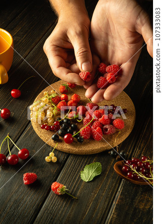 Many different fresh berries on the kitchen table. Man's hands sorting raspberries before preparing fruit drink or compote 107333083