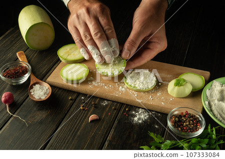 The chef dusts the sliced zucchini with flour before roasting. The concept of cooking vegetable marrow on the kitchen table with vegetables and spices The chef dusts the sliced zucchini with flour before roasting. The concept of cooking vegetable marrow on the kitchen table with vegetables and spices 107333084