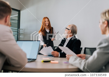 Remote view of cheerful female colleague come in for business briefing and take seat at table with mature adult business partner. Business team start working at desk in office, discussing strategy. 107338549