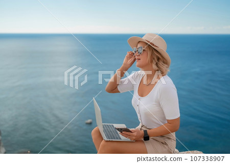 Freelance women sea working on the computer. Good looking middle aged woman typing on a laptop keyboard outdoors with a beautiful sea view. The concept of remote work. 107338907