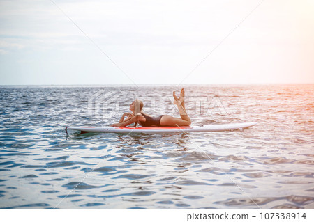 Woman sup sea. Sports girl on a surfboard in the sea on a sunny summer day. In a black bathing suit, he sits on a sapa in the sea. Rest on the sea. Woman sup sea. Sports girl on a surfboard in the sea on a sunny summer day. In a black bathing suit, he sits on a sapa in the sea. Rest on the sea. 107338914