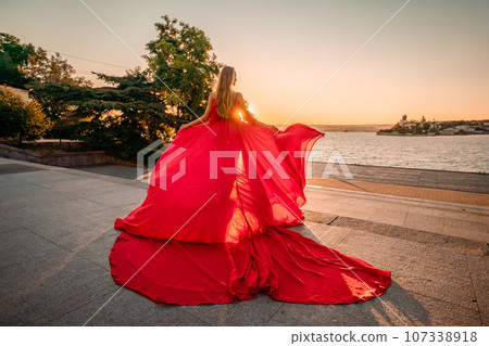 Sunrise red dress. A woman in a long red dress against the backdrop of sunrise, bright golden light of the sun's rays. The concept of femininity, harmony. 107338918