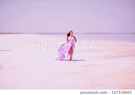 Woman pink salt lake. Against the backdrop of a pink salt lake, a woman in a long pink dress takes a leisurely stroll along the white, salty shore, capturing a wanderlust moment. 107339065