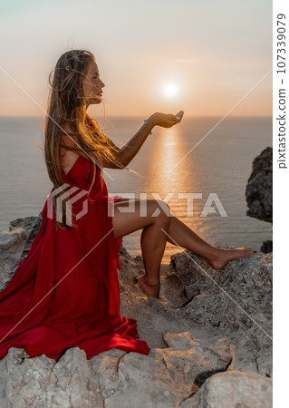 Woman sunset sea red dress, side view a happy beautiful sensual woman in a red long dress posing on a rock high above the sea on sunset. Woman sunset sea red dress, side view a happy beautiful sensual woman in a red long dress posing on a rock high above the sea on sunset. 107339079