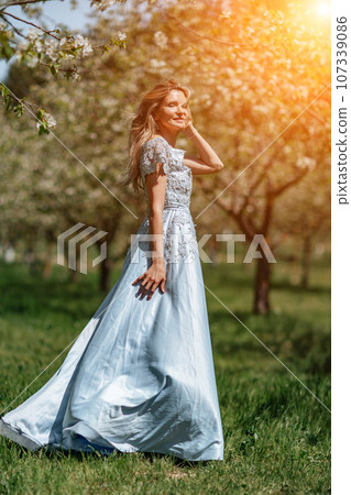 Blond garden. Portrait of a blonde in the park. Happy woman with long blond hair in a blue dress. 107339086