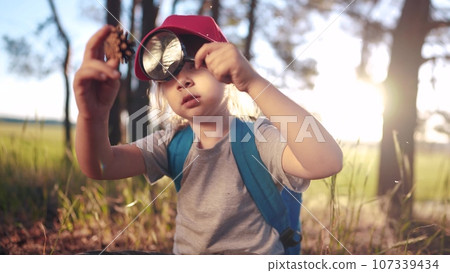 child girl boy scout studying a pine cone in the forest with a lifestyle magnifying glass. happy family child dream concept. child tourist explorer with magnifying glass in the forest child girl boy scout studying a pine cone in the forest with a lifestyle magnifying glass. happy family child dream concept. child tourist explorer with magnifying glass in the forest 107339434