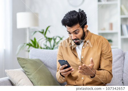 Man received bad news message with sad news online while sitting on couch in living room using app on phone. 107342657