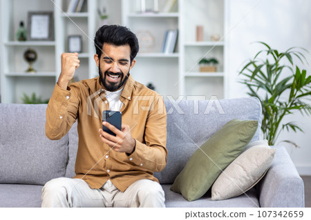 Young man received an online notification of a winning message sitting at home in the living room on the sofa, the winner is satisfied with results of the achievement joyfully holding his hand up. 107342659