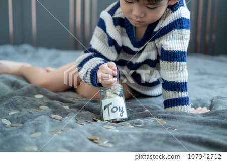 Asian boy sits and puts coins in a glass jar to save money to buy toys. 107342712