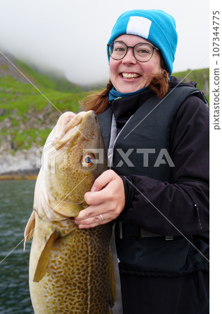 Happy young woman holding big arctic cod. Norway happy fishing. Fisherwoman with cod fish in hands Happy young woman holding big arctic cod. Norway happy fishing. Fisherwoman with cod fish in hands 107344775