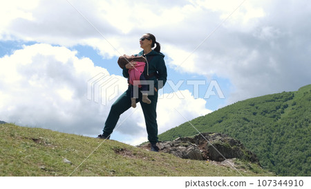 Cheerful young mother hiker with sleepy baby enjoying view while hiking in mountains 107344910