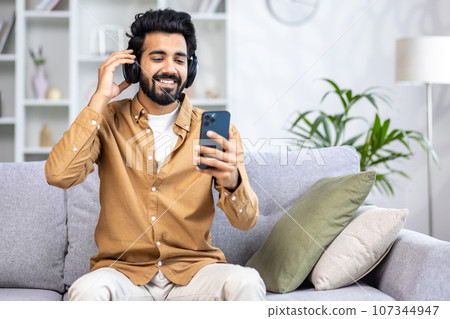 Young joyful man sitting at home on sofa in living room, using app on phone and headphones listening to music and online radio streaming. 107344947