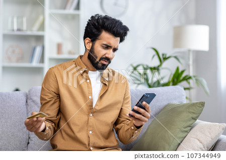 Cheated and sad man received a rejected money transfer error sitting on sofa in living room, holding bank credit card and phone, negative account balance. Cheated and sad man received a rejected money transfer error sitting on sofa in living room, holding bank credit card and phone, negative account balance. 107344949