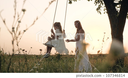 Happy mother rides little girl with teddy bear on swings in park at sunset. Little girl plays swings with mother in evening field. Happy family of mother and daughter play swings in summer park 107346814