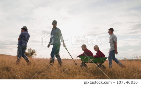 Mother carries baby and father pulls brothers in cart walking with family along field at sunset. Children with parents enjoy family weekend in farm field. Farmer family with children rest in field 107346816