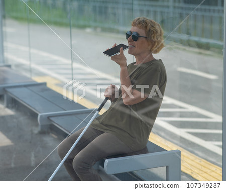 Elderly blind woman sitting at a bus stop and using a smartphone. Elderly blind woman sitting at a bus stop and using a smartphone. 107349287