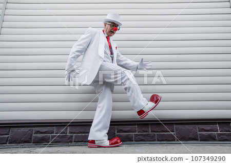 An elderly man in a white suit, huge boots and a clown nose walks funny. An elderly man in a white suit, huge boots and a clown nose walks funny. 107349290