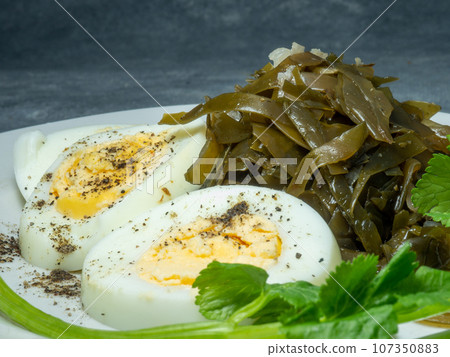 Seaweed salad and cut boiled egg. Dish on the table in a plate. Peppered half of an egg. Parsley greens as decoration. Seaweed salad and cut boiled egg. Dish on the table in a plate. Peppered half of an egg. Parsley greens as decoration. 107350883