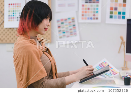 Young graphic designer using a tablet and stylus pen while sitting at her desk in office. Young graphic designer using a tablet and stylus pen while sitting at her desk in office. 107352526