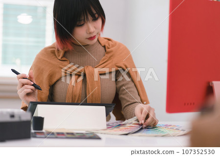 A creative and stylish young female freelance designer is using a tablet and stylus pen while sitting at her desk in office. 107352530