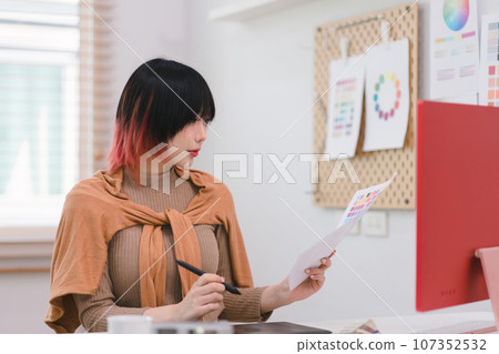 A creative and stylish young female freelance designer is using a tablet and stylus pen while sitting at her desk in office. 107352532