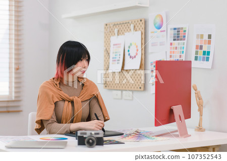 A creative and stylish young female freelance designer is using a tablet and stylus pen while sitting at her desk in her home office. 107352543