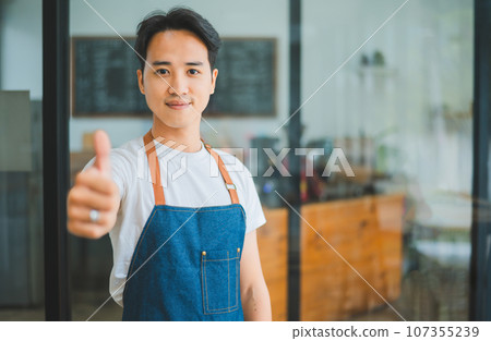 Smiling Asian coffee shop owner standing in front of cashier counter.welcome customers who come in to place orders in the cafe. Smiling Asian coffee shop owner standing in front of cashier counter.welcome customers who come in to place orders in the cafe. 107355239
