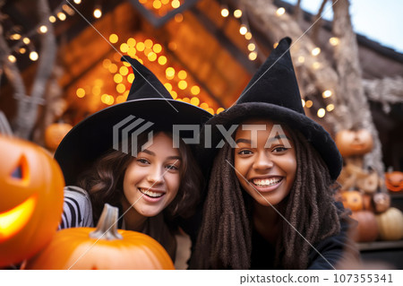 Two multiracial young women friends wearing witch hats having fun and take selfies at Halloween Two multiracial young women friends wearing witch hats having fun and take selfies at Halloween 107355341