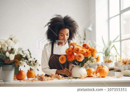 Attractive african american woman florist working in flower shop. Creation of autumn decor for Halloween celebration. 107355351
