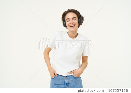 Portrait of young female model, wearing headphones, listens to music, smiling and laughing, standing over white background Portrait of young female model, wearing headphones, listens to music, smiling and laughing, standing over white background 107357156