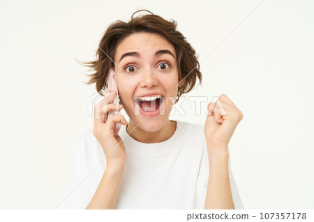 Portrait of amazed brunette girl looks surprised while talks on mobile phone, answers a call, amazed by big news, gasping shocked, standing over white background Portrait of amazed brunette girl looks surprised while talks on mobile phone, answers a call, amazed by big news, gasping shocked, standing over white background 107357178