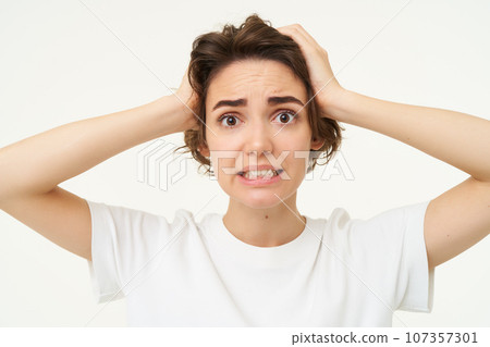 Close up of woman in panic, holding hands on head and clenching teeth, looking troubled, puzzled and frustrated by something, standing over white background Close up of woman in panic, holding hands on head and clenching teeth, looking troubled, puzzled and frustrated by something, standing over white background 107357301