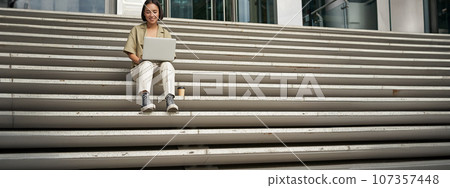 Asian girl student sits on stairs near campus, types on laptop, does her homework outdoors Asian girl student sits on stairs near campus, types on laptop, does her homework outdoors 107357448