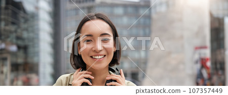 Vertical shot of beautiful asian woman posing with headphones around neck, smiling and laughing, standing on street in daylight 107357449