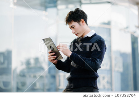 Young Asian business man working at outside business center with laptop, tablet, smartphone and taking notes on the paper. Young Asian business man working at outside business center with laptop, tablet, smartphone and taking notes on the paper. 107358704