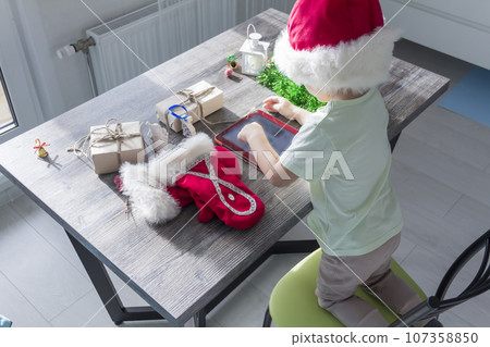 A small child in a Santa Claus hat, with Christmas gifts, is sitting at a New Year's table with a tablet in his hands. 107358850
