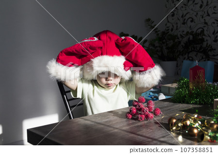 A small child in a Santa Claus hat and red mittens with Christmas gifts is sitting at the New Year's table. A small child in a Santa Claus hat and red mittens with Christmas gifts is sitting at the New Year's table. 107358851
