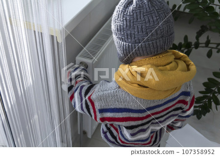 A small child warmly dressed in a sweater and a hat turns the tap on the radiator. The concept of crisis and lack of heating in homes. A small child warmly dressed in a sweater and a hat turns the tap on the radiator. The concept of crisis and lack of heating in homes. 107358952