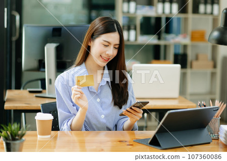 Asian Woman using smart phone for mobile payments online shopping,omni channel,sitting on table,virtual icons graphics interface screen in morning light. 107360086