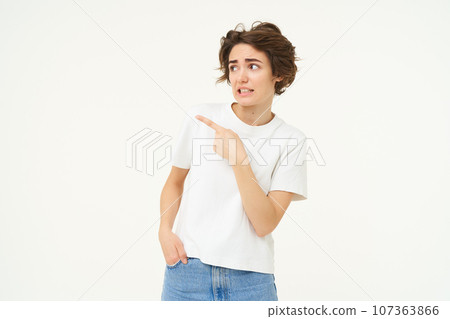Portrait of woman looking with doubt, pointing left, staring at something with unsure, concerned face expression, standing over white studio background Portrait of woman looking with doubt, pointing left, staring at something with unsure, concerned face expression, standing over white studio background 107363866