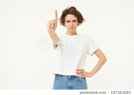 Portrait of serious woman shows one finger, rule or taboo gesture, disapprove something, scolding person, frowning at camera, looking angry, standing over white background Portrait of serious woman shows one finger, rule or taboo gesture, disapprove something, scolding person, frowning at camera, looking angry, standing over white background 107363883