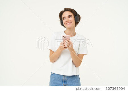 Image of happy young girl listens to music in headphones, standing with smartphone, using mobile phone streaming app, white background 107363904