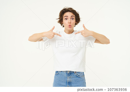 Image of girl with surprised look, pointing at herself with amazed face, being chosen or winning something, standing over white background 107363936