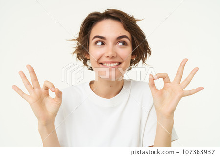 Image of excited brunette woman, smiles, shows okay, ok gesture, looks amused and happy, stands over white studio background 107363937