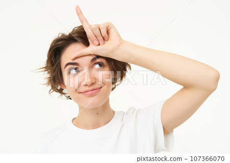 Close up of woman shows loser gesture, l letter on forehead and smiling, mocking, makes fun of someone, standing over white background Close up of woman shows loser gesture, l letter on forehead and smiling, mocking, makes fun of someone, standing over white background 107366070