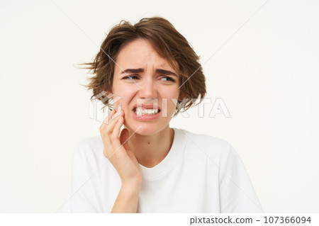 Close up of woman has a toothache, touches her teeth and frowns from painful discomfort, stands over white background Close up of woman has a toothache, touches her teeth and frowns from painful discomfort, stands over white background 107366094